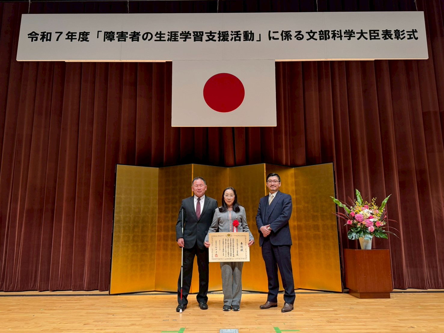 令和７年度「障害者の生涯学習支援活動」に係る文部科学大臣表彰式の横断幕の前で表彰状を持って立つ西村専務、梶本会長、辻副会長