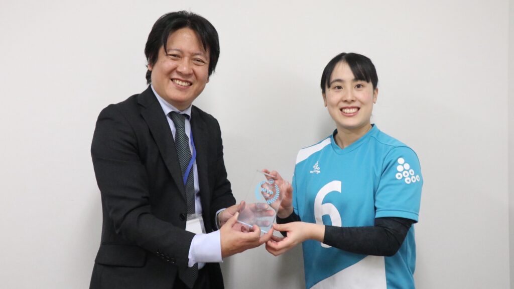 ＦＰパートナー徳山様からトロフィーを受け取る高橋利恵子選手。徳山様は黒いスーツ。高橋選手はメルヴェイユのユニフォームを着用している。